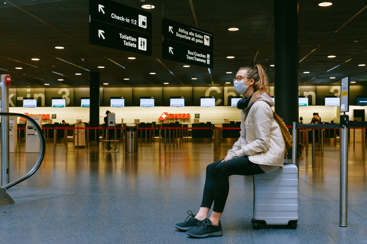 A woman in a mask sitting on a suitcase in an airport representing mandatory masks when travelling