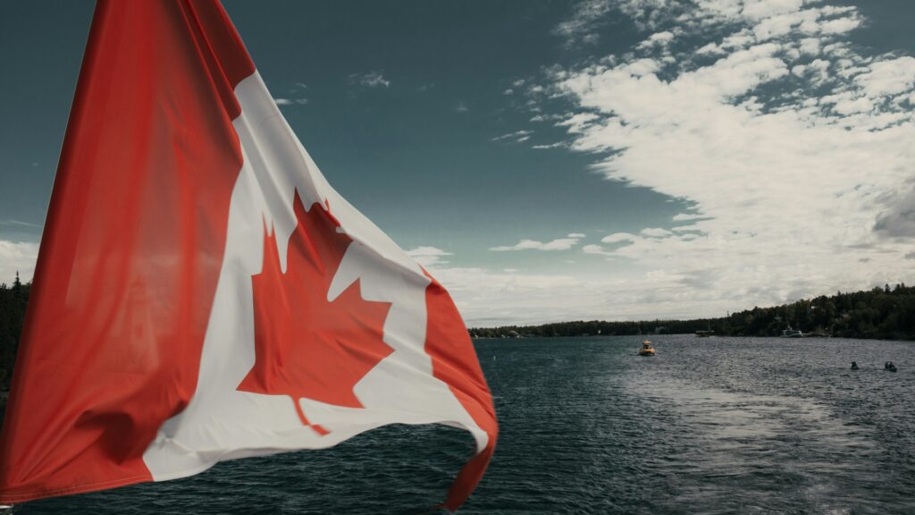A Canadian flag billows in the foreground, with a serene lake and tree-lined shore in the background under a cloudy sky, visually representing the national context of the blog discussing Canada's 2025-2026 immigration plan and its impact on pathways to permanent residency, international students, and workers.