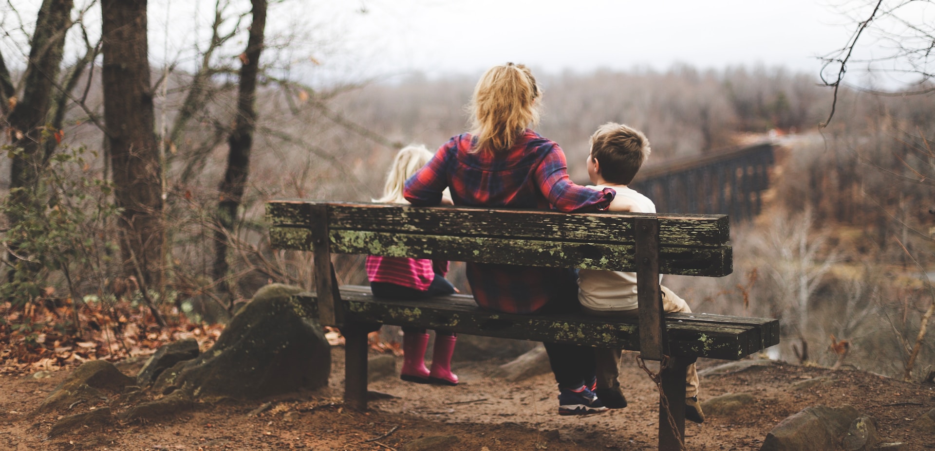 A mother and her children look our over a forested ravine with a railway bridge stretching arcoss it from a wooden bench in late fall, representing Citizenship by Descent in Canada.